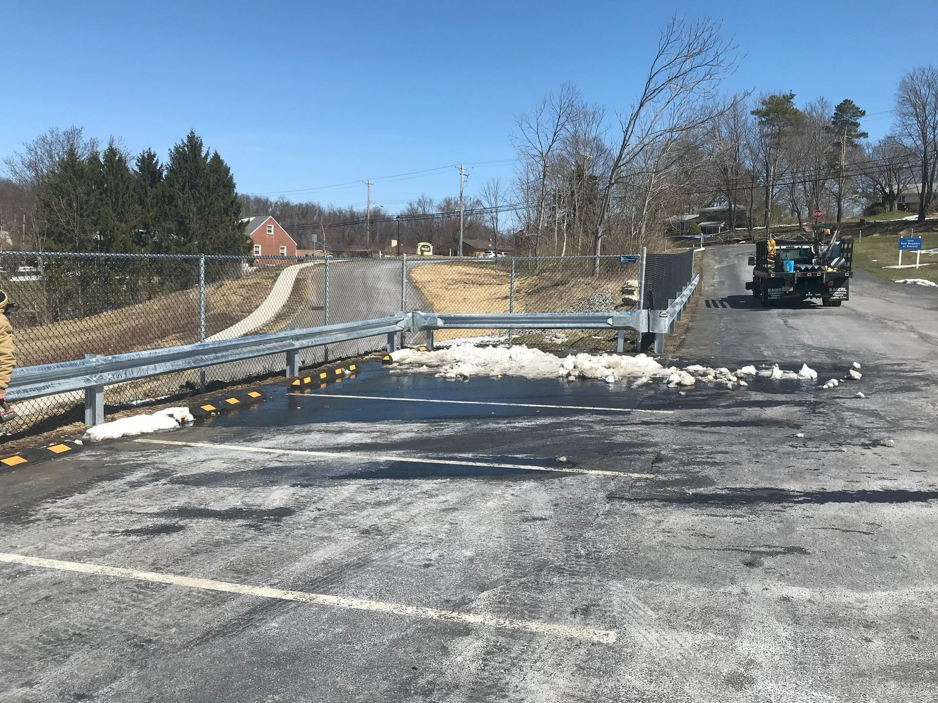 Snowy roadside construction area with a blue truck, metal guardrail, and leafless trees under a clear sky