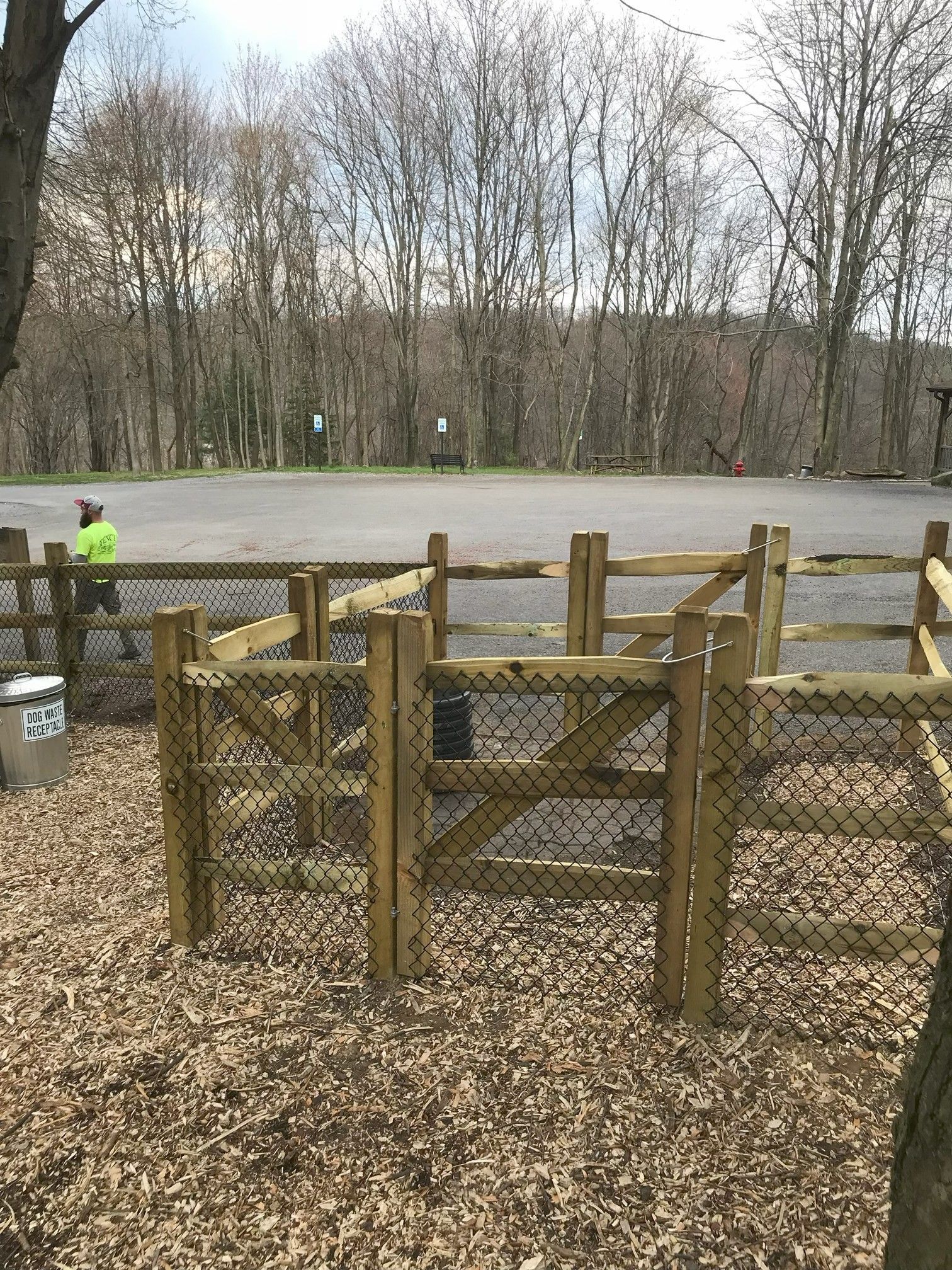 Wooded park with fenced gravel area and a worker in a neon jacket near a paved path
