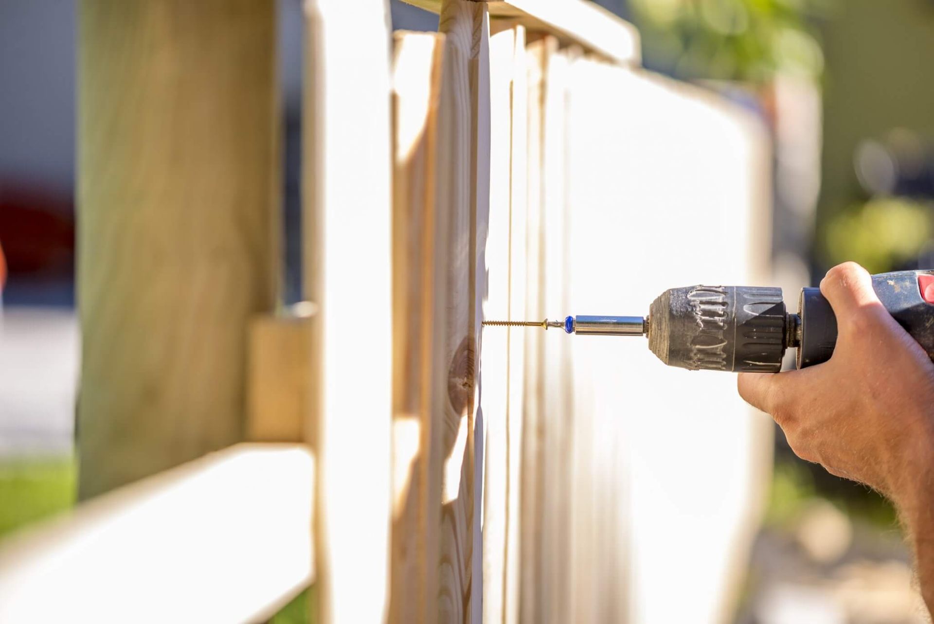 Hand drilling a screw into a light-colored wooden fence in bright sunlight
