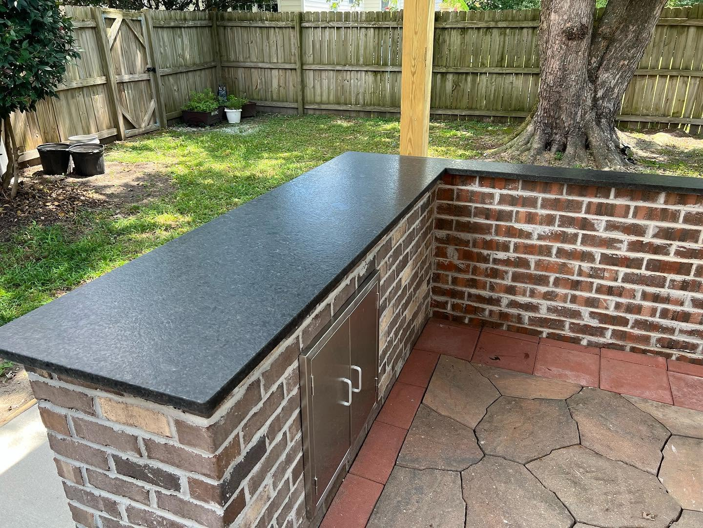 Brick outdoor kitchen counter with black granite top in a fenced backyard, beside a tree and patio stones