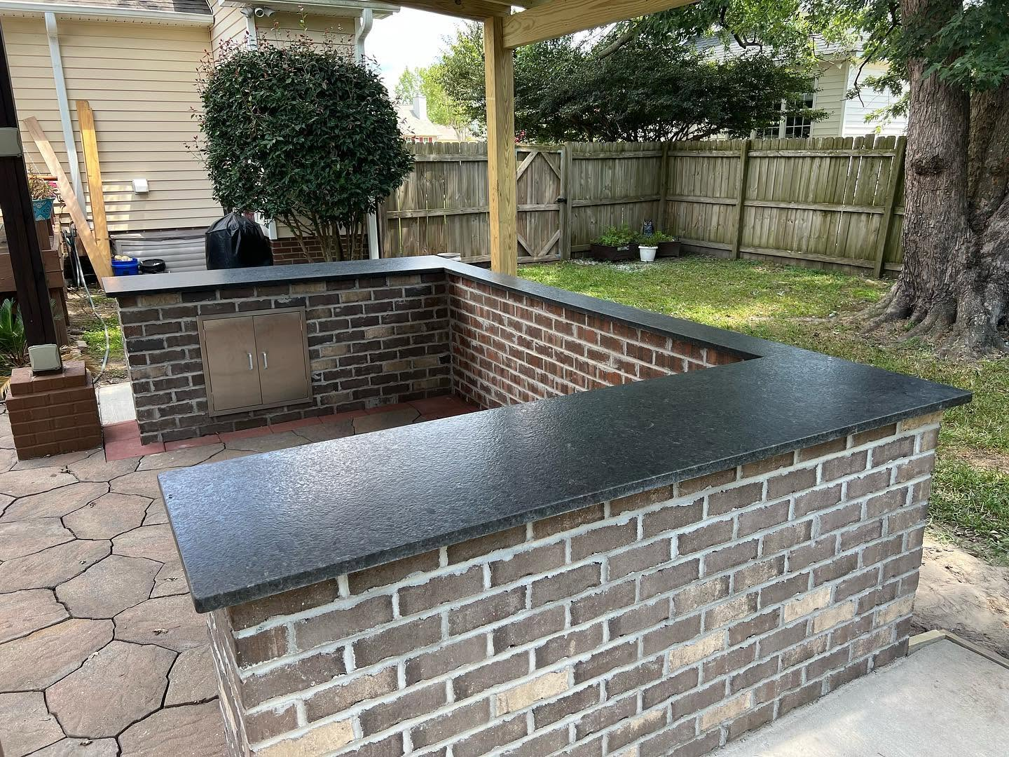 Outdoor brick bar with dark stone countertop under a wooden pergola in a backyard
