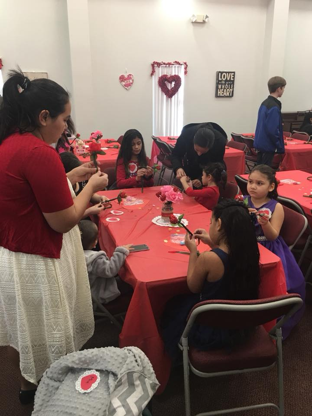 A woman in a red shirt is standing in front of a group of children sitting at tables