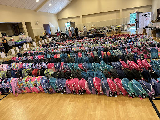 A large amount of backpacks are lined up on a wooden floor in a gym.
