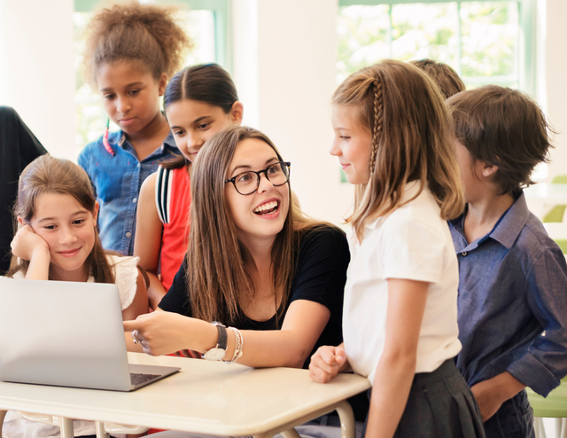 A group of children are listening to their female tutor as she explains something on her laptop.