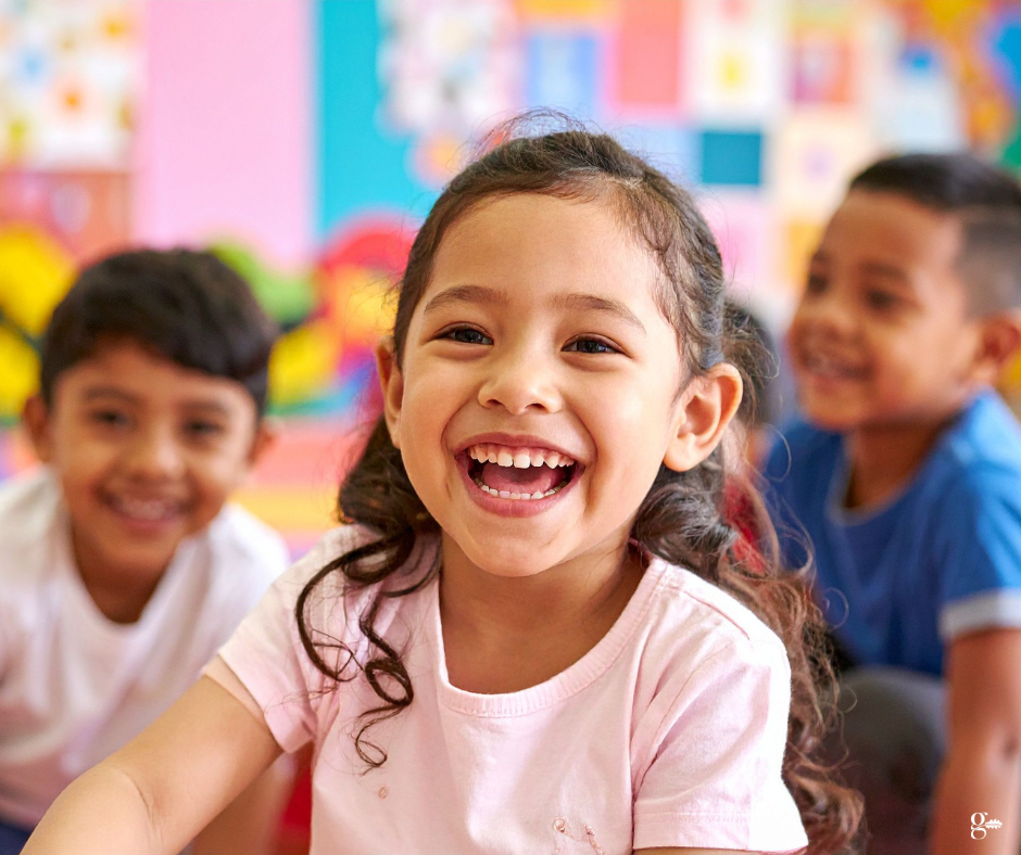 Smiling child in a pink shirt with two other children smiling in a brightly coloured classroom.