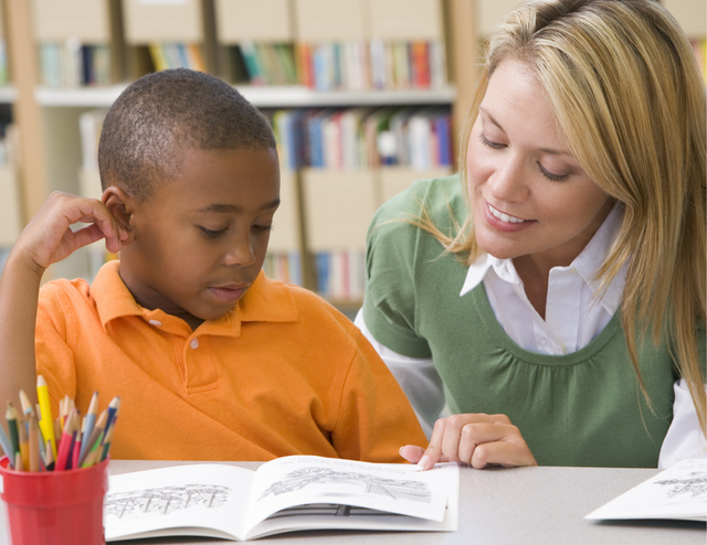 A little boy is sitting reading a book with a female tutor.