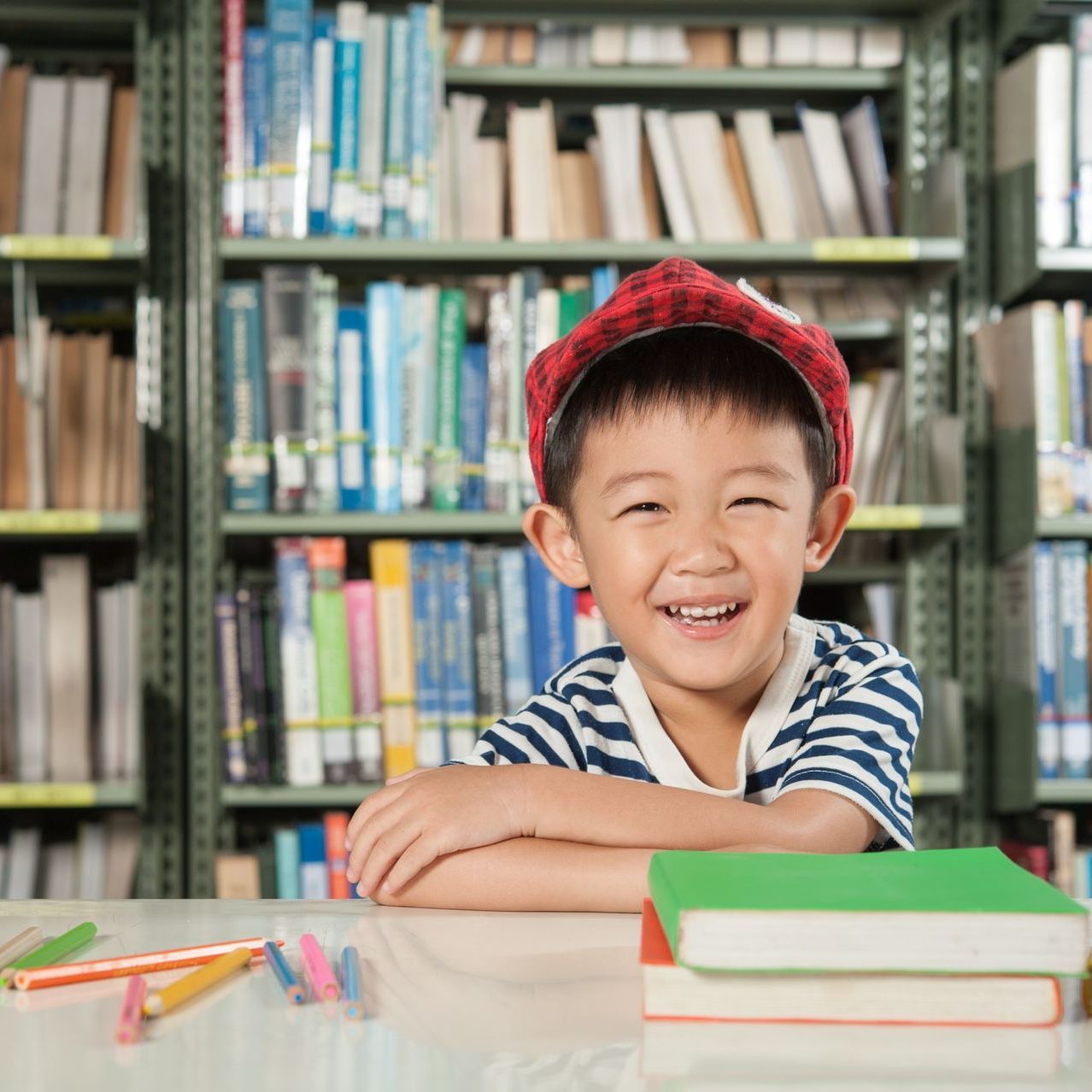A young boy is sitting at a table in a library with books and pencils.