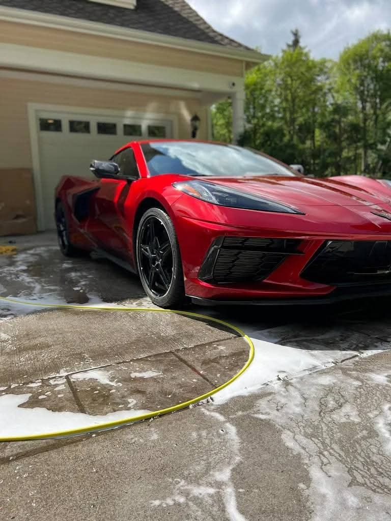 Red sports car parked in a driveway beside a house, with wet pavement and a garage door in the background