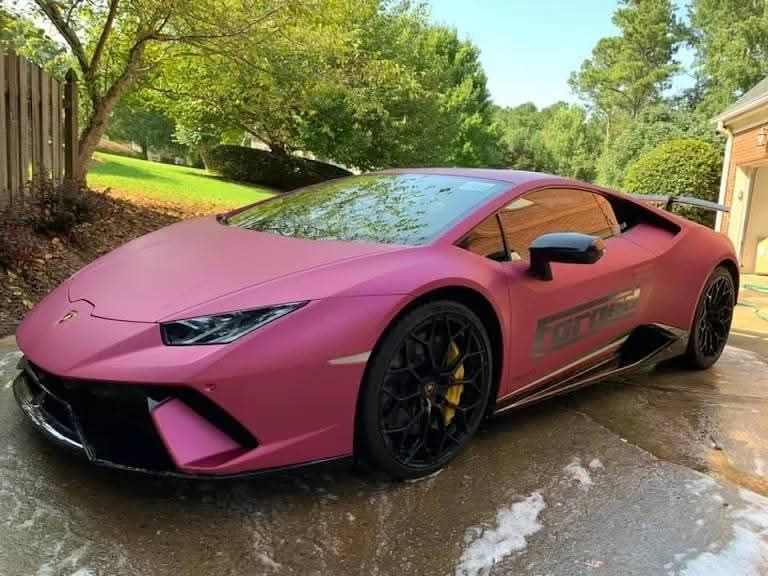 Pink sports car parked on a wet driveway beside a house, with trees and greenery around.