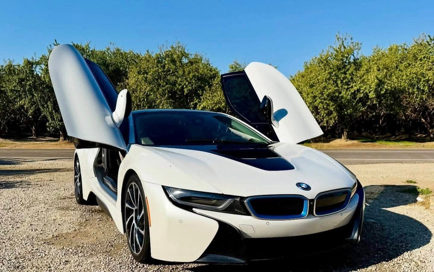 White BMW i8 with butterfly doors open, parked on a road with trees in the background.