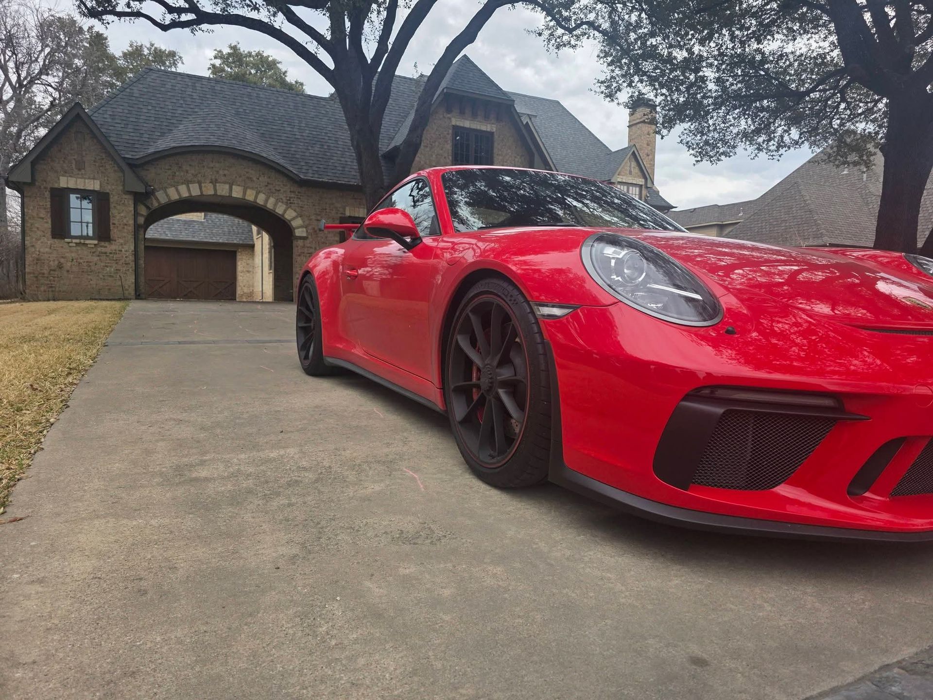 Red sports car parked in a driveway outside a brick house with trees overhead