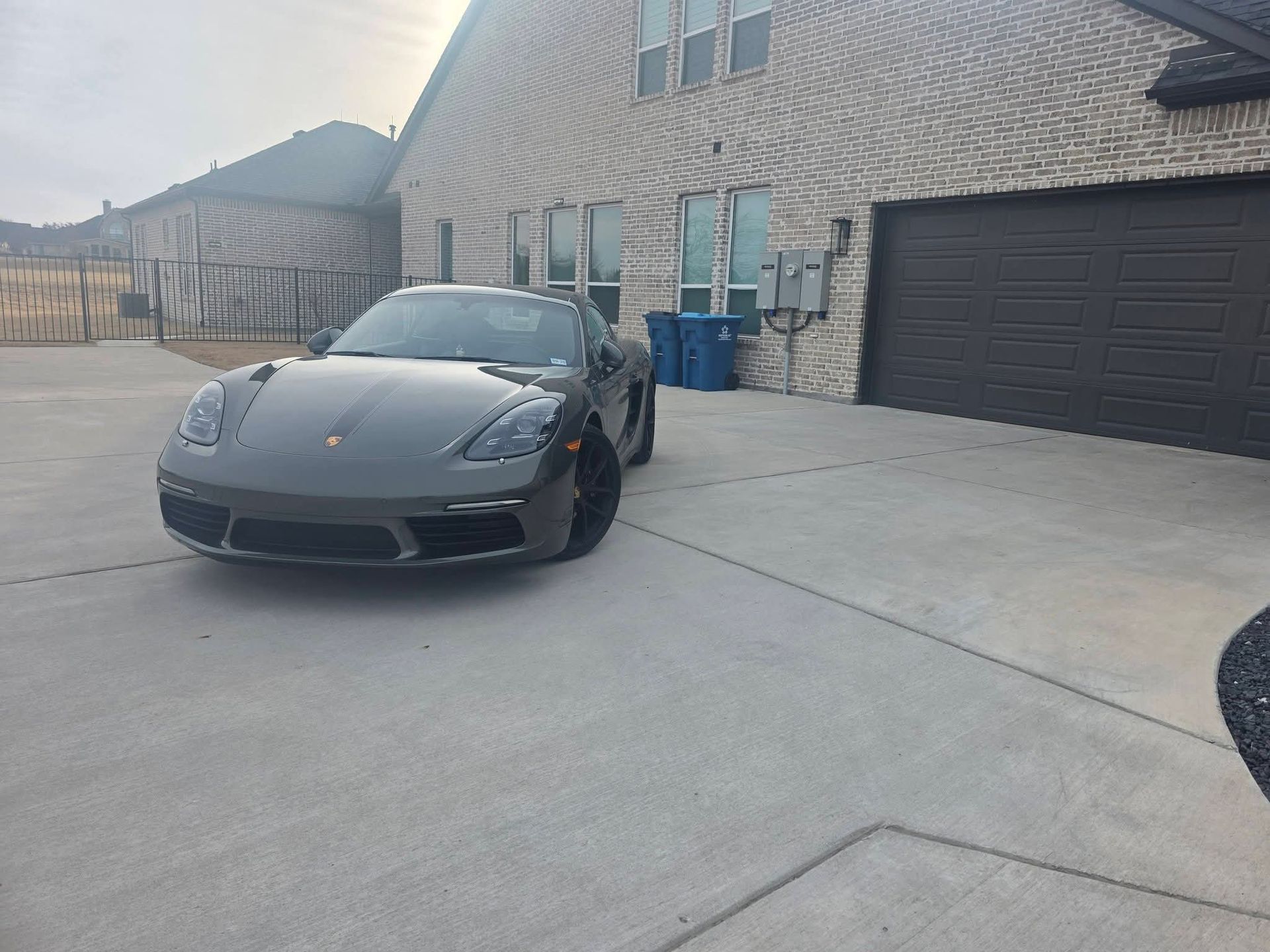 Dark gray sports car parked in a driveway beside a brick house and closed garage door