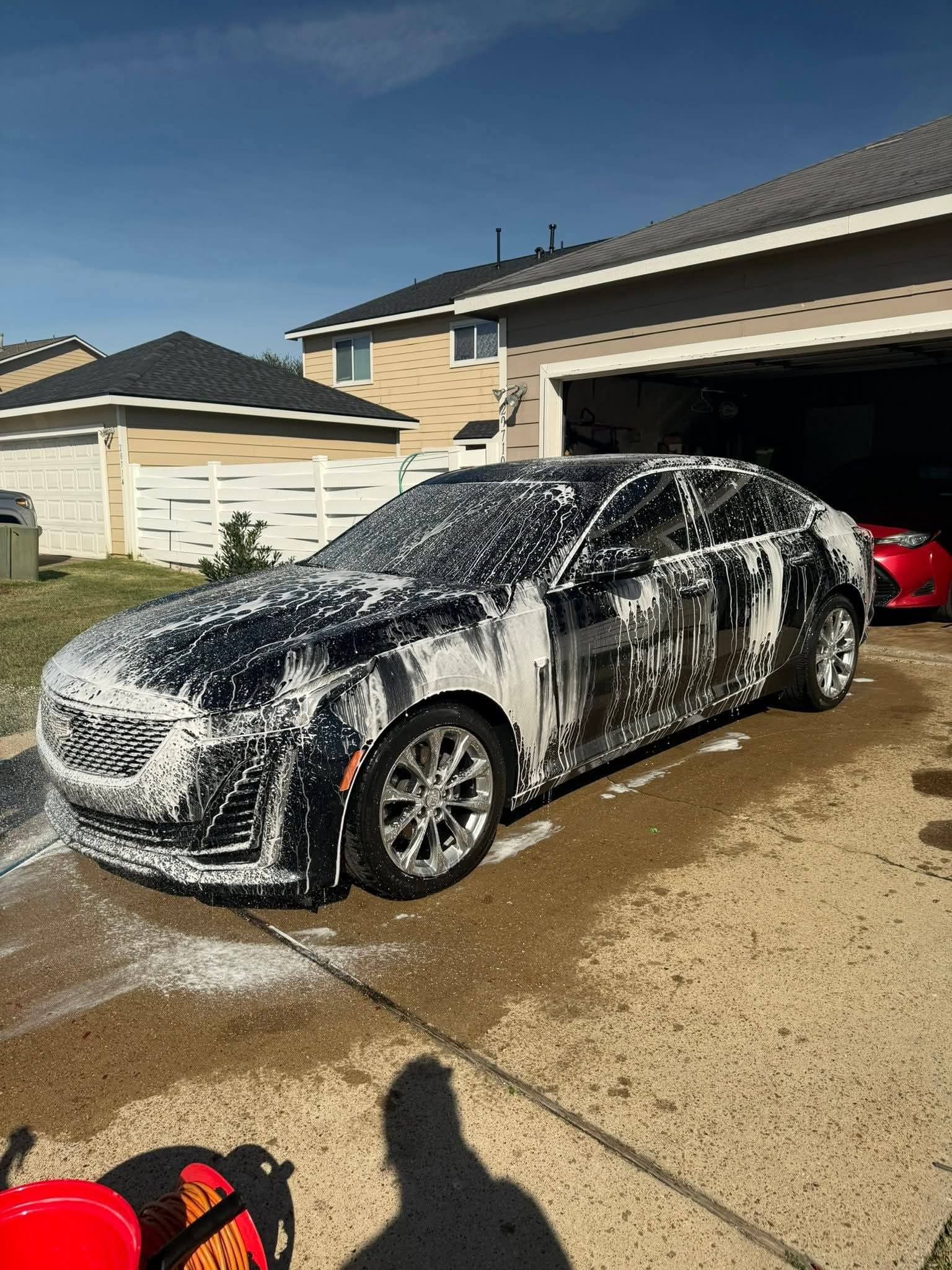 Foam-covered black sedan parked in a driveway beside a garage, with a red car inside.