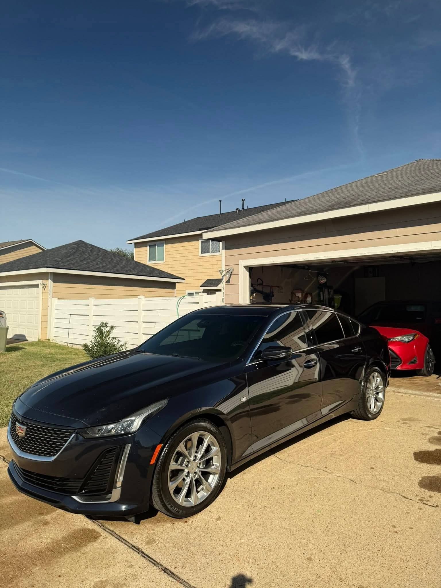 Black convertible sports car parked in a driveway beside a house, with a red car in the garage.
