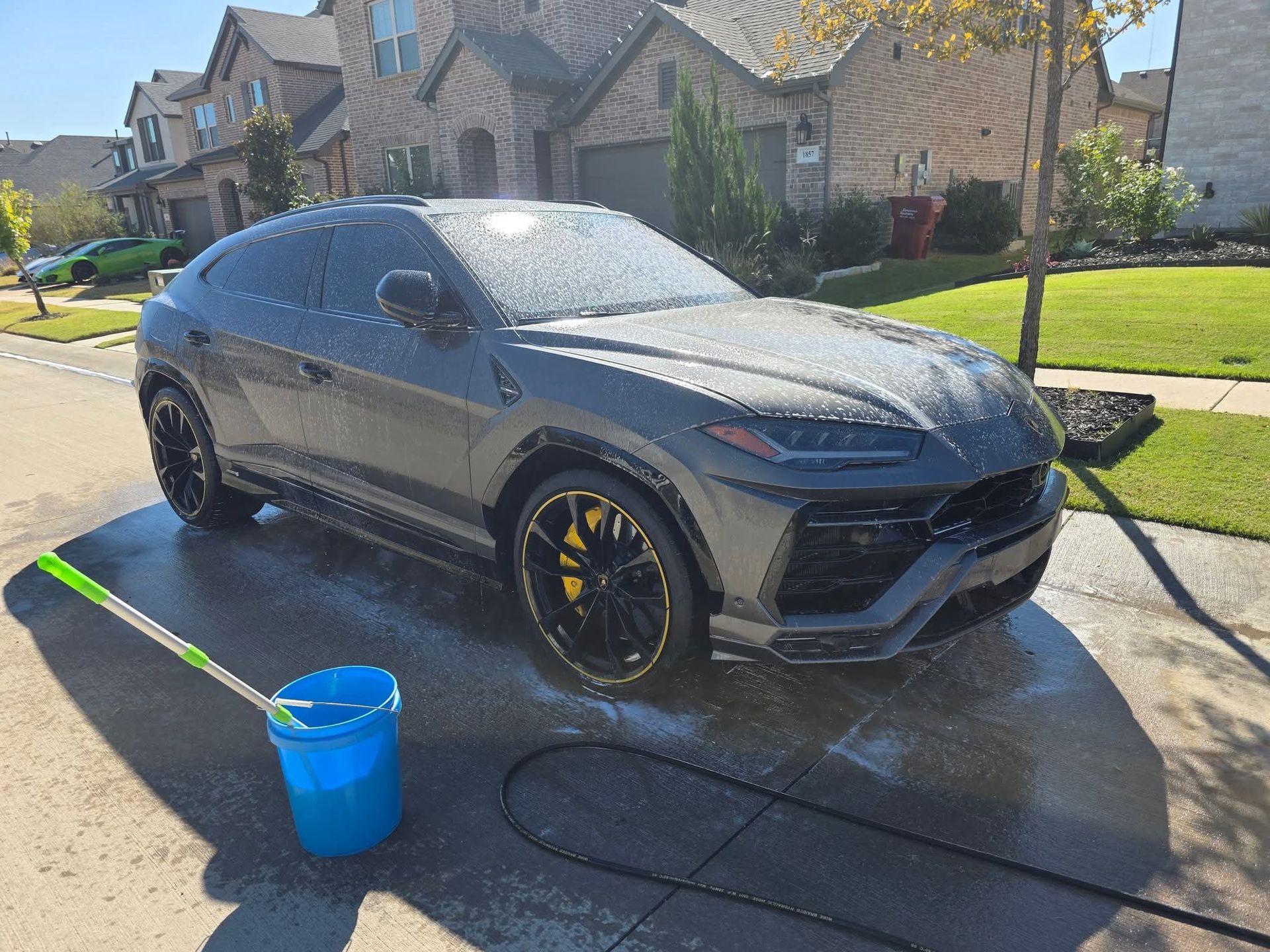 Black SUV being washed in a driveway with a blue bucket and hose on a sunny suburban street