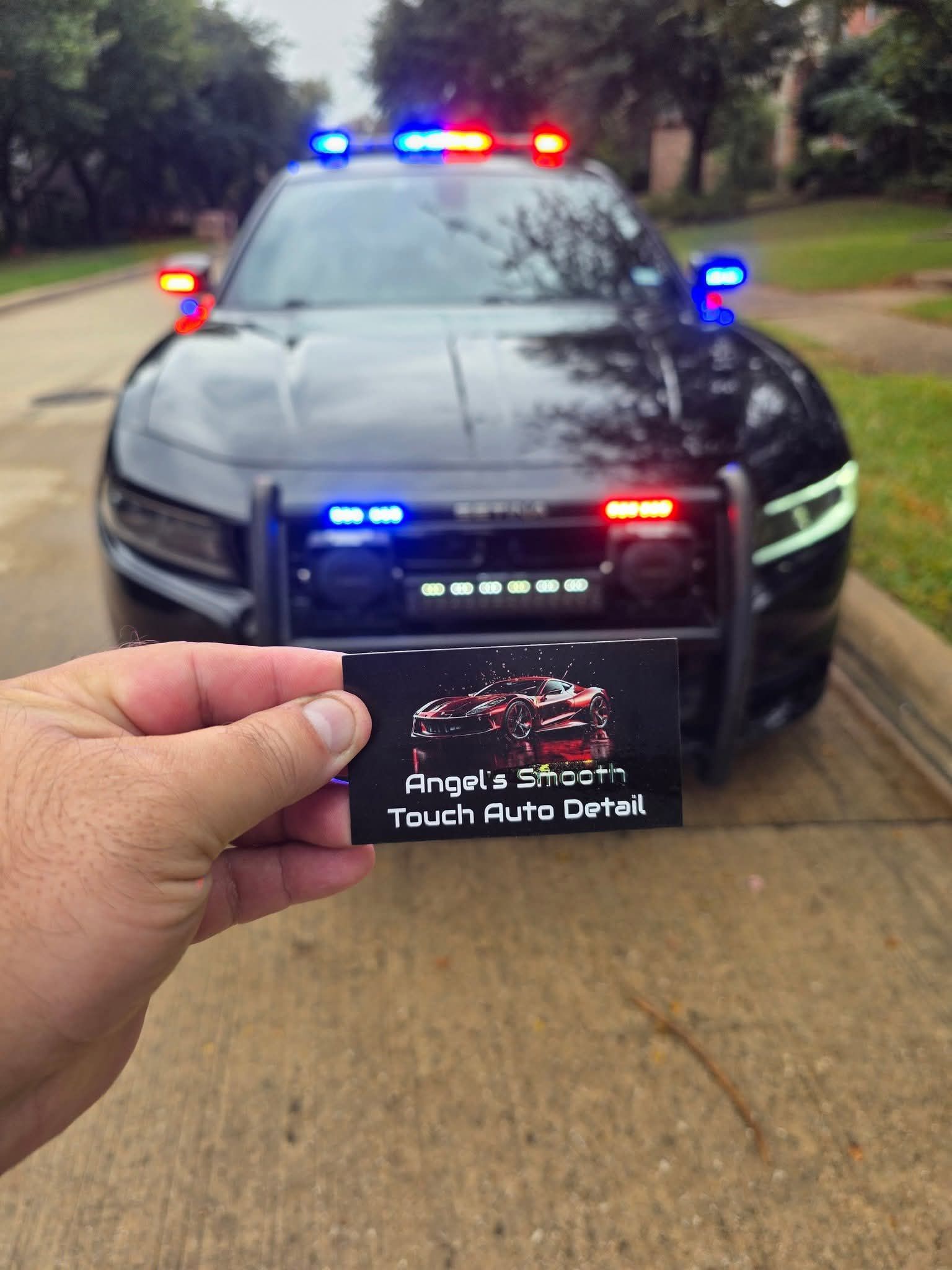 Hand holding a business card in front of a black police-style car with flashing red and blue lights on a driveway