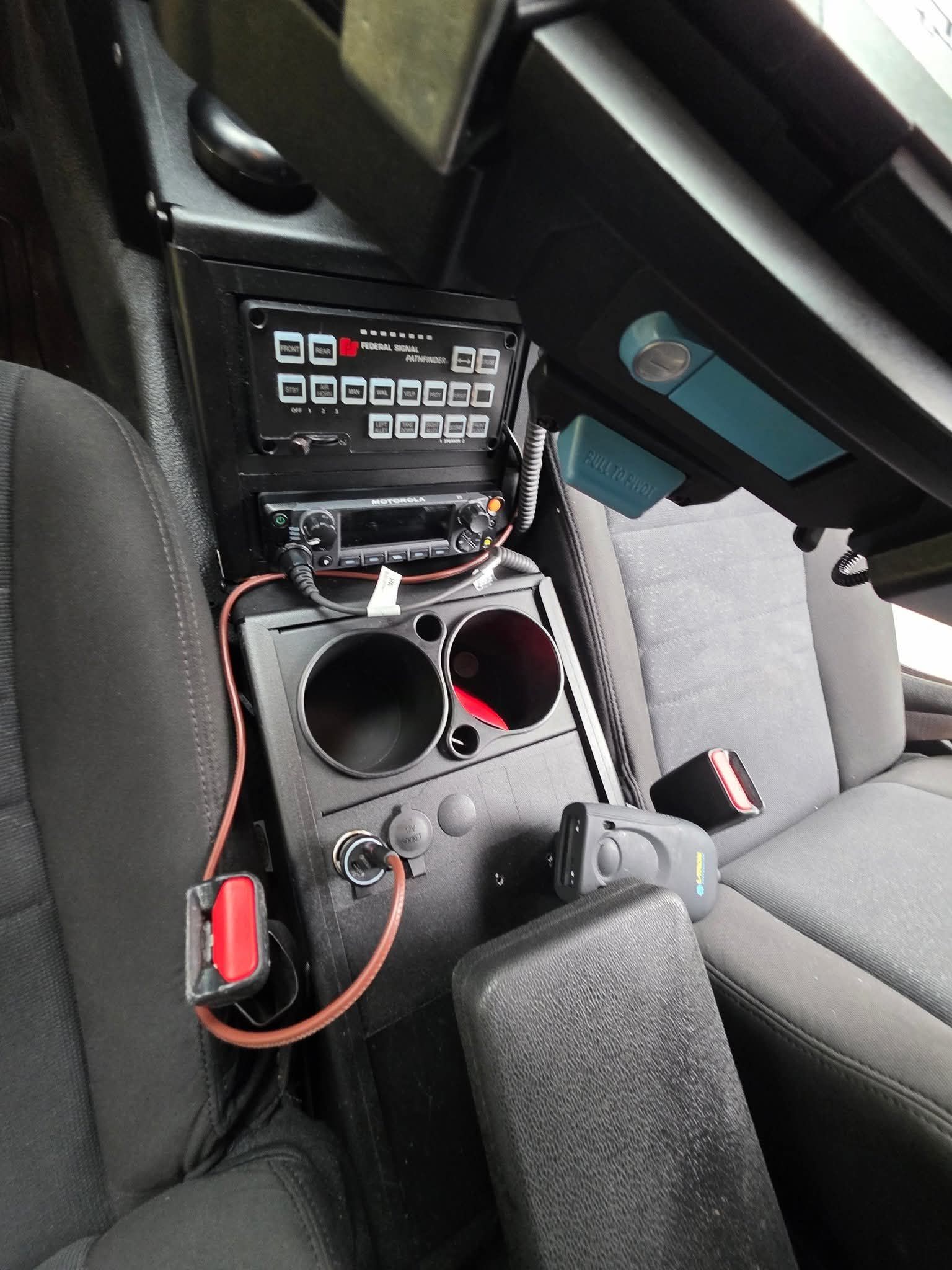 Car center console with radio, cup holders, and exposed wiring beside gray seats
