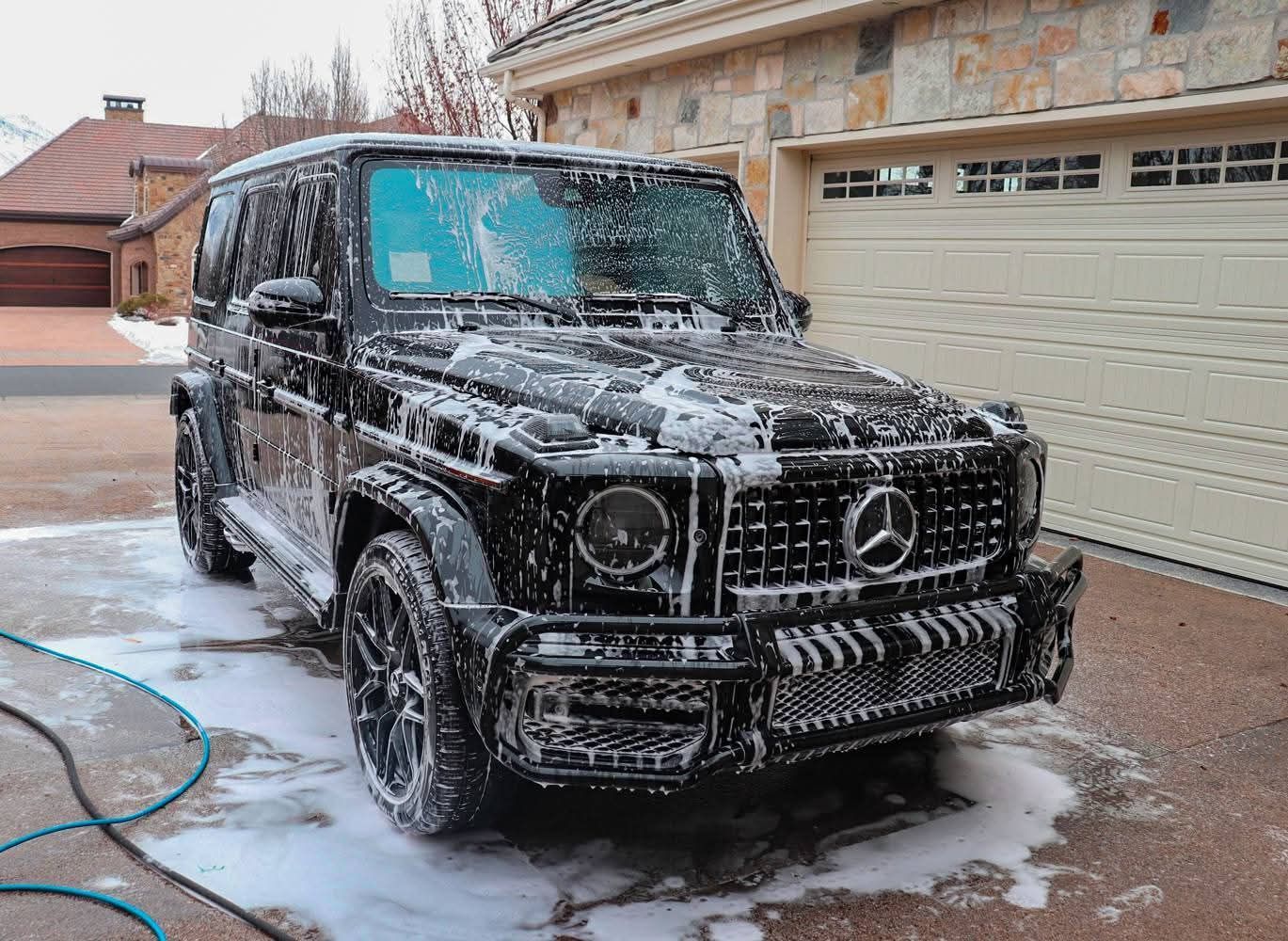 Black Mercedes SUV being washed with soap foam in a driveway beside a garage.