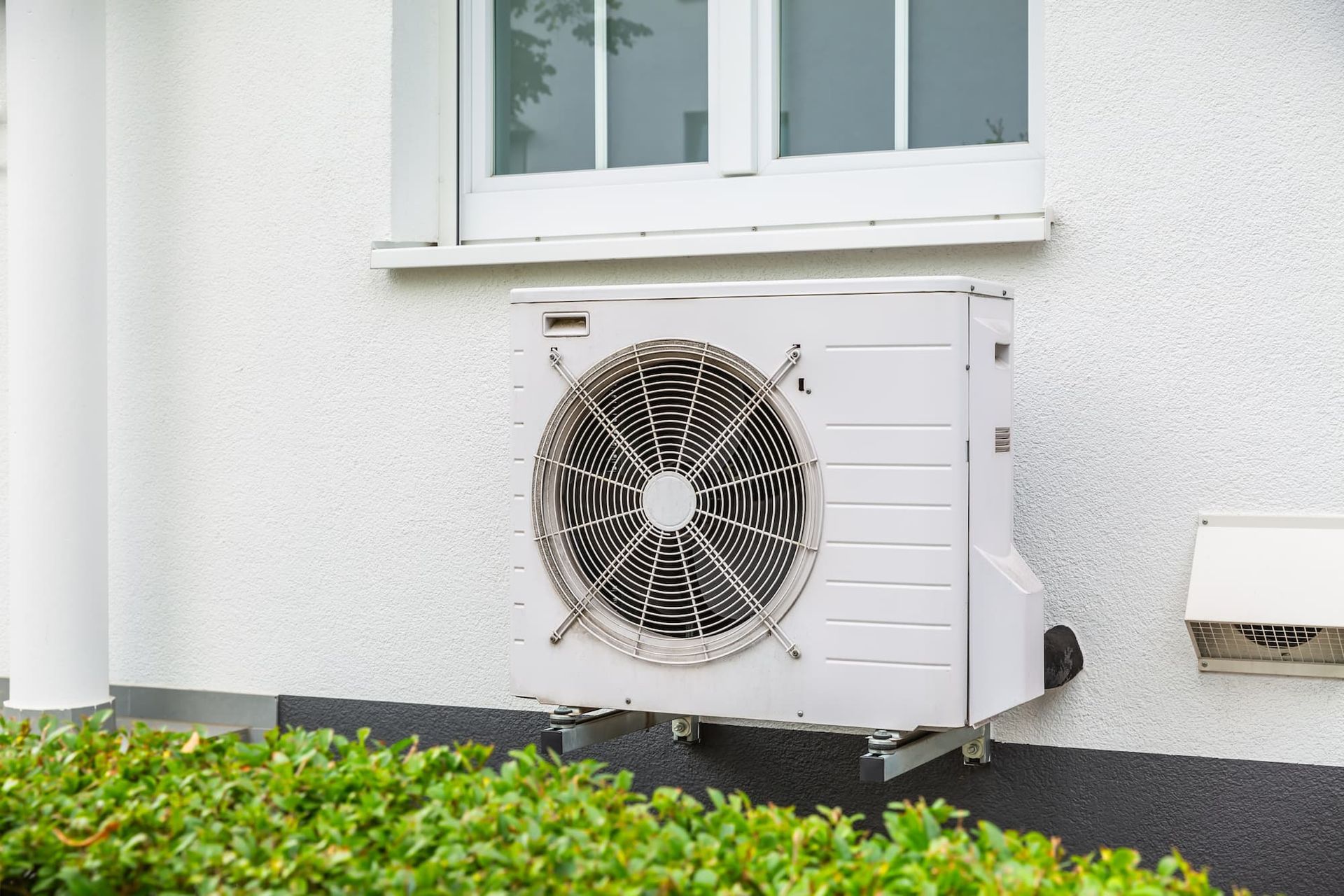 Air conditioner unit mounted on a white house exterior beneath a window, with greenery in the foreground.