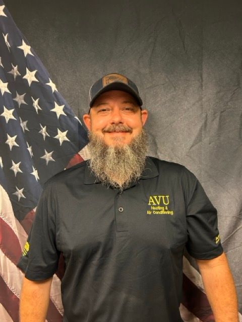 Man with beard and hat in black polo shirt stands in front of American flag.