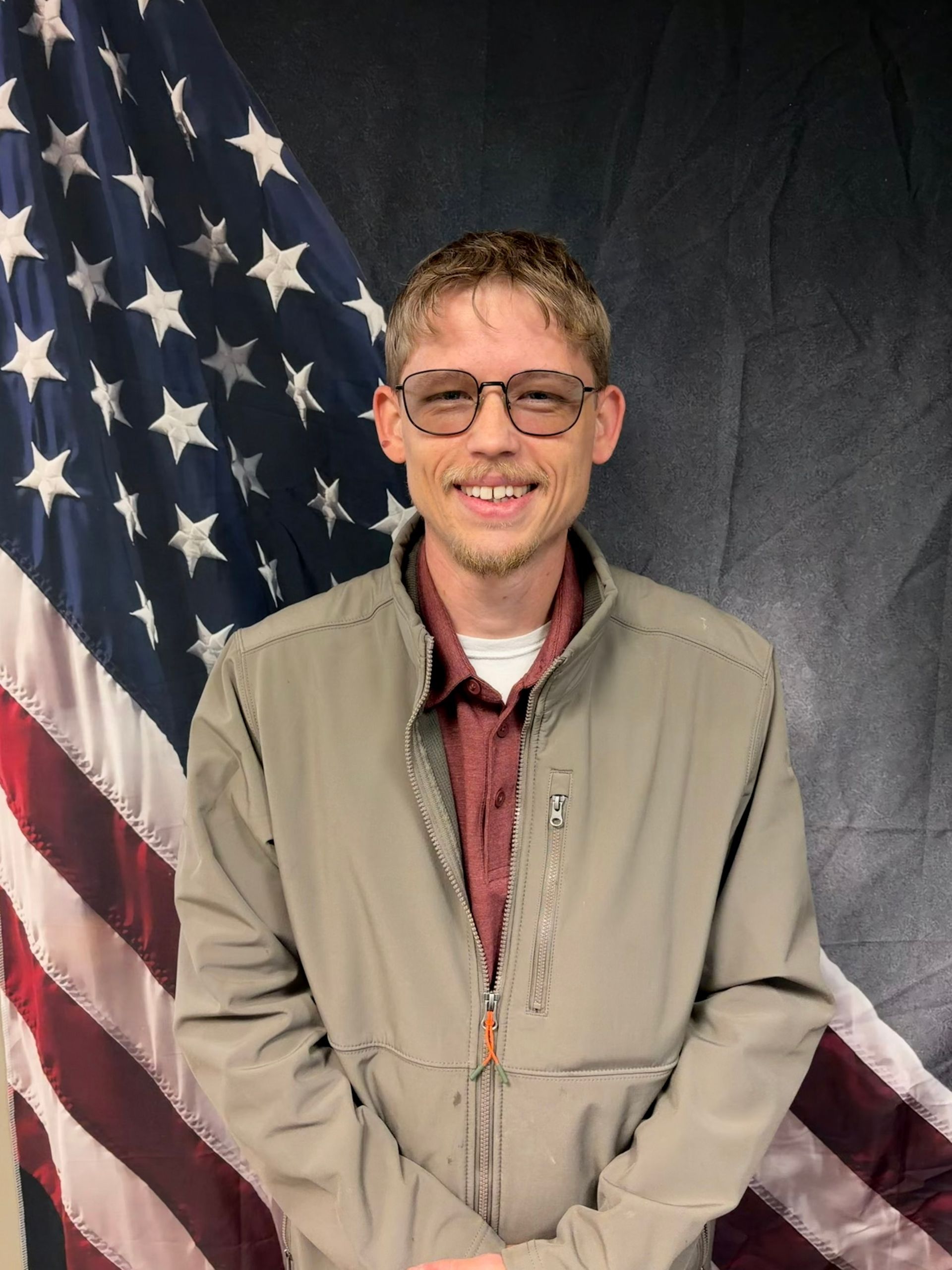 Man in glasses and jacket smiling, standing in front of American flag.
