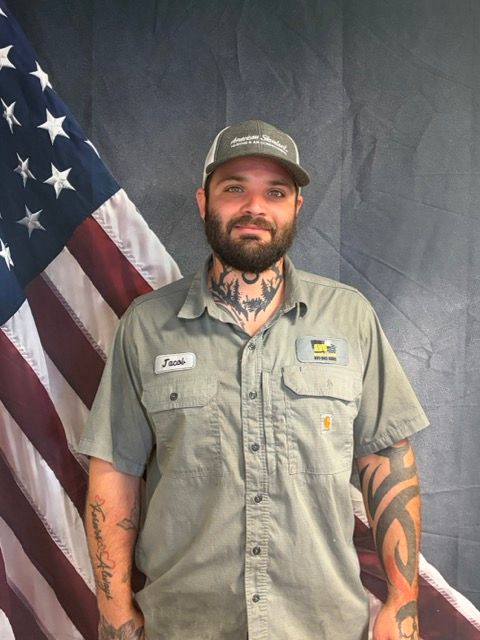 Man with beard wearing cap and work shirt, standing in front of an American flag.