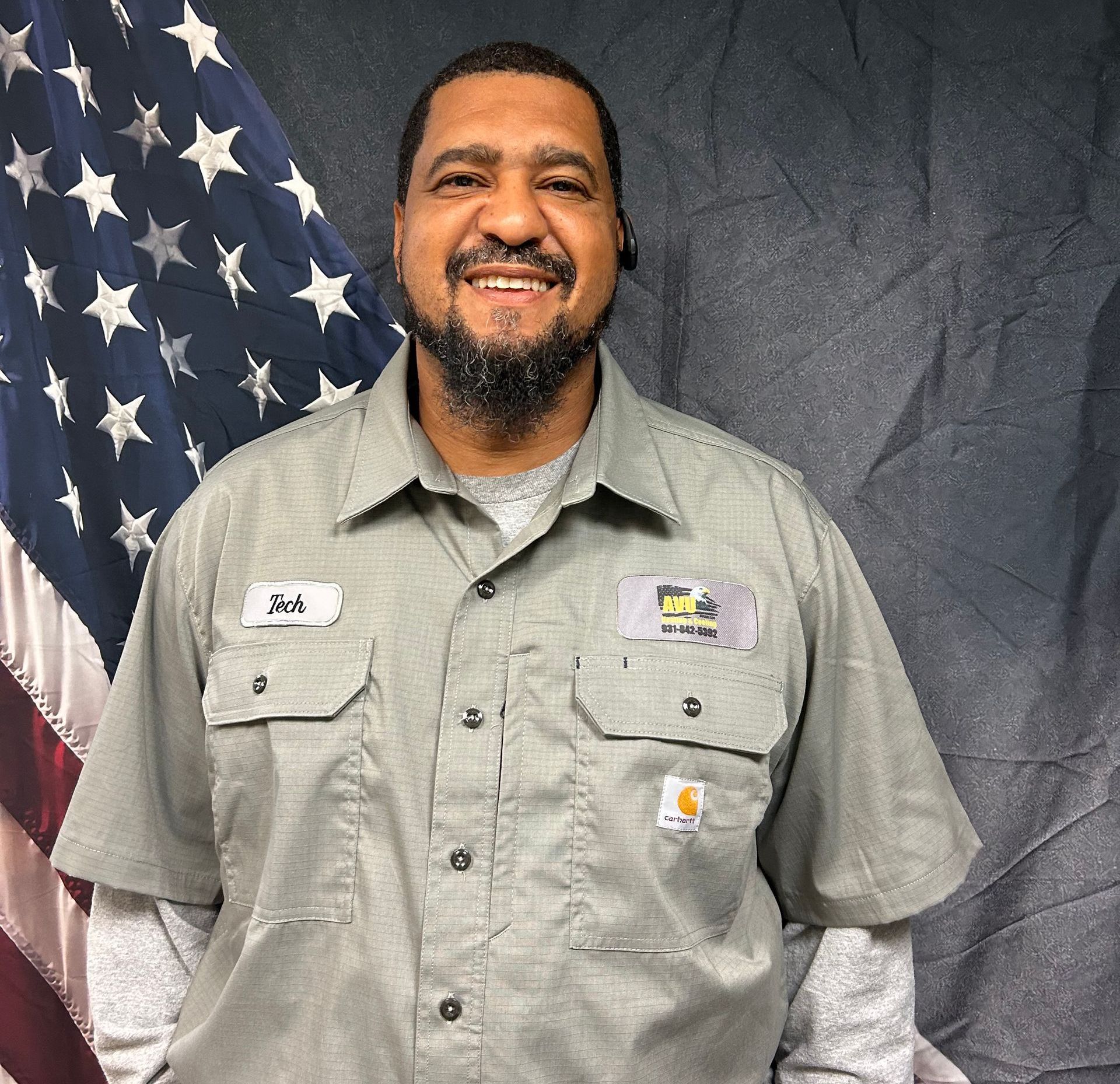 Man in work shirt smiles in front of American flag, logo on shirt, wearing a headset.