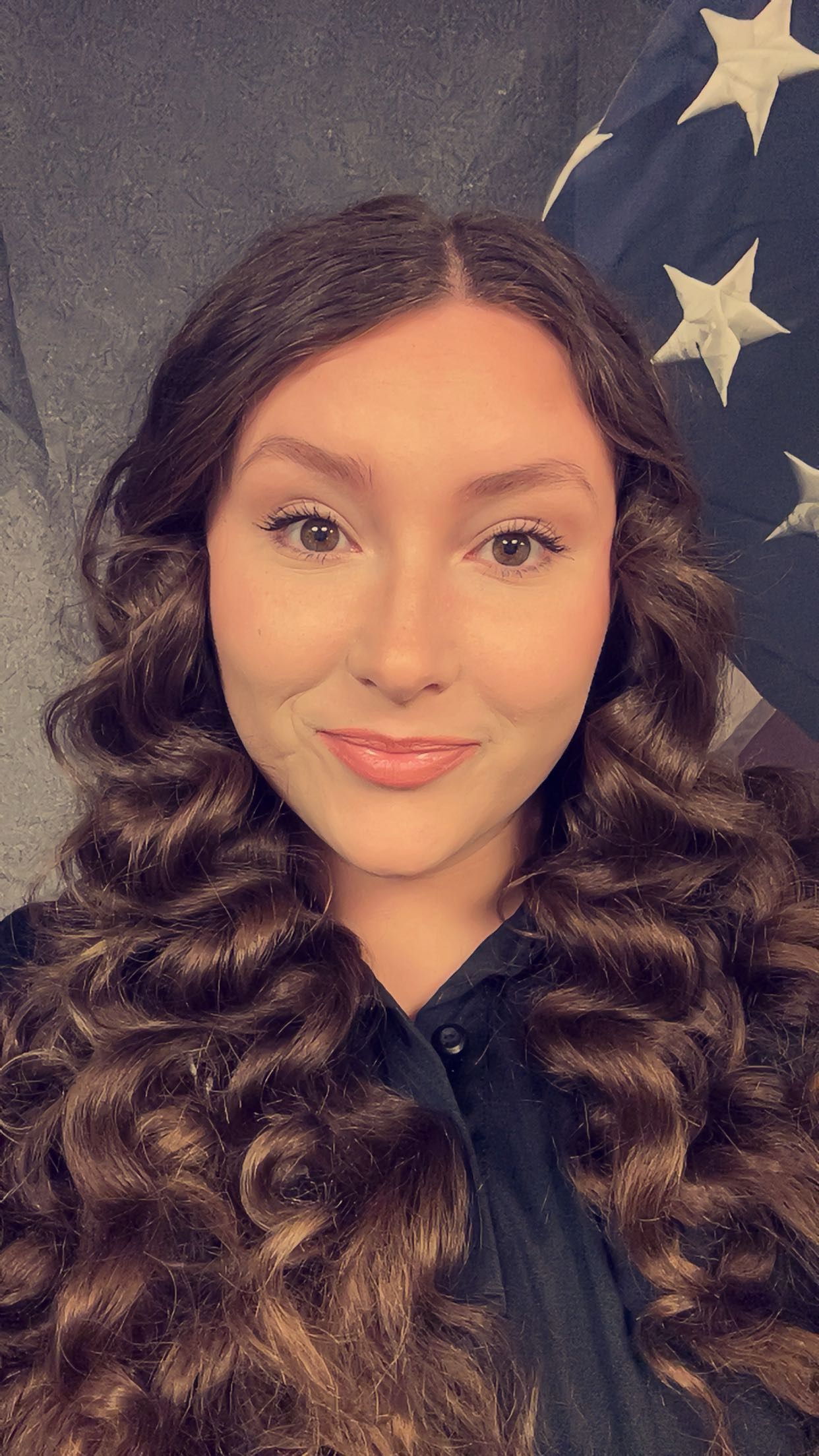 A woman with long curly hair is taking a selfie in front of an american flag.