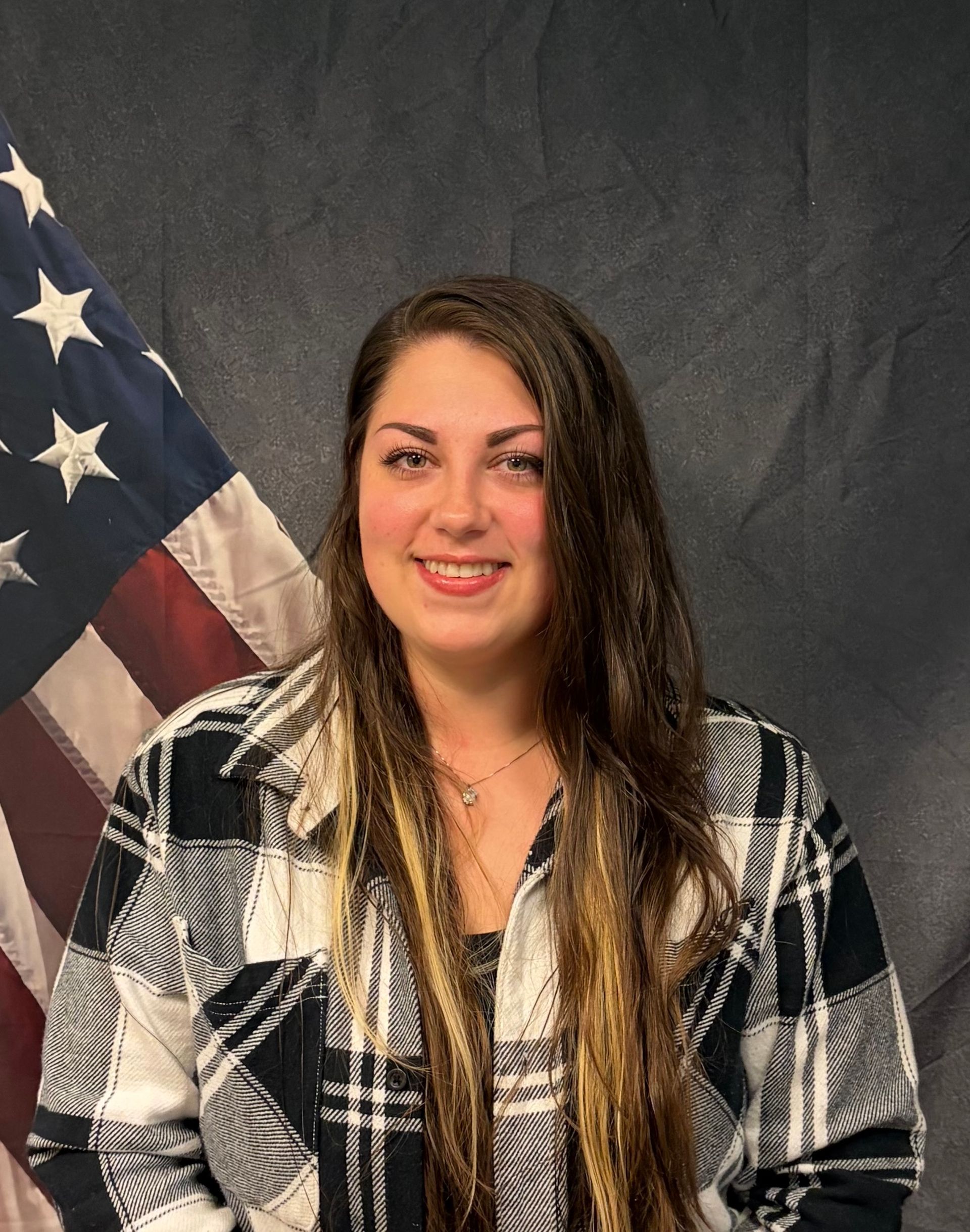 Woman smiling, wearing a black and white plaid jacket, in front of an American flag.