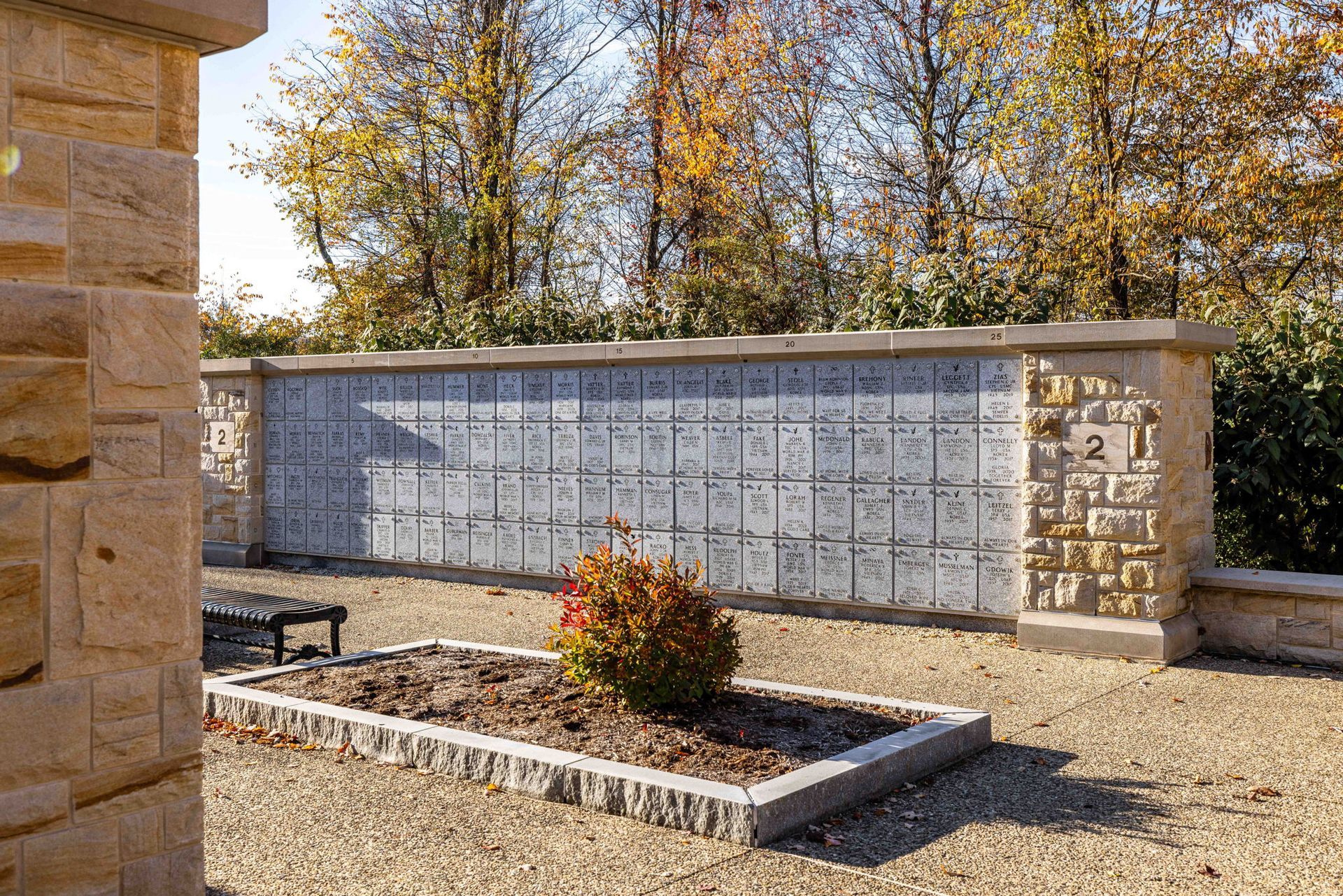Outdoor memorial wall with engraved plaques and a small flower bed in a courtyard, autumn trees behind