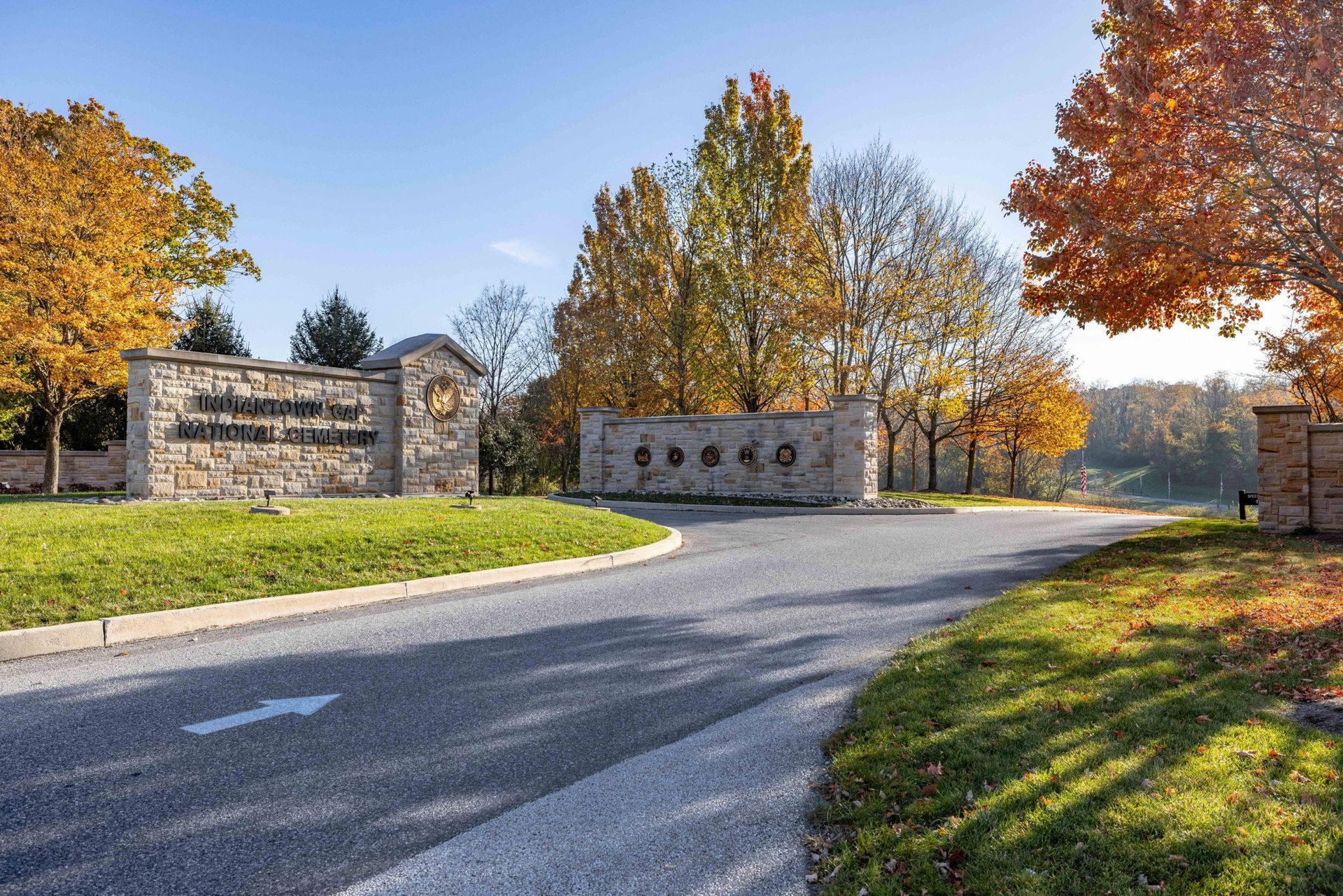 Stone entrance gate beside a curving road, framed by autumn trees with yellow and orange leaves.