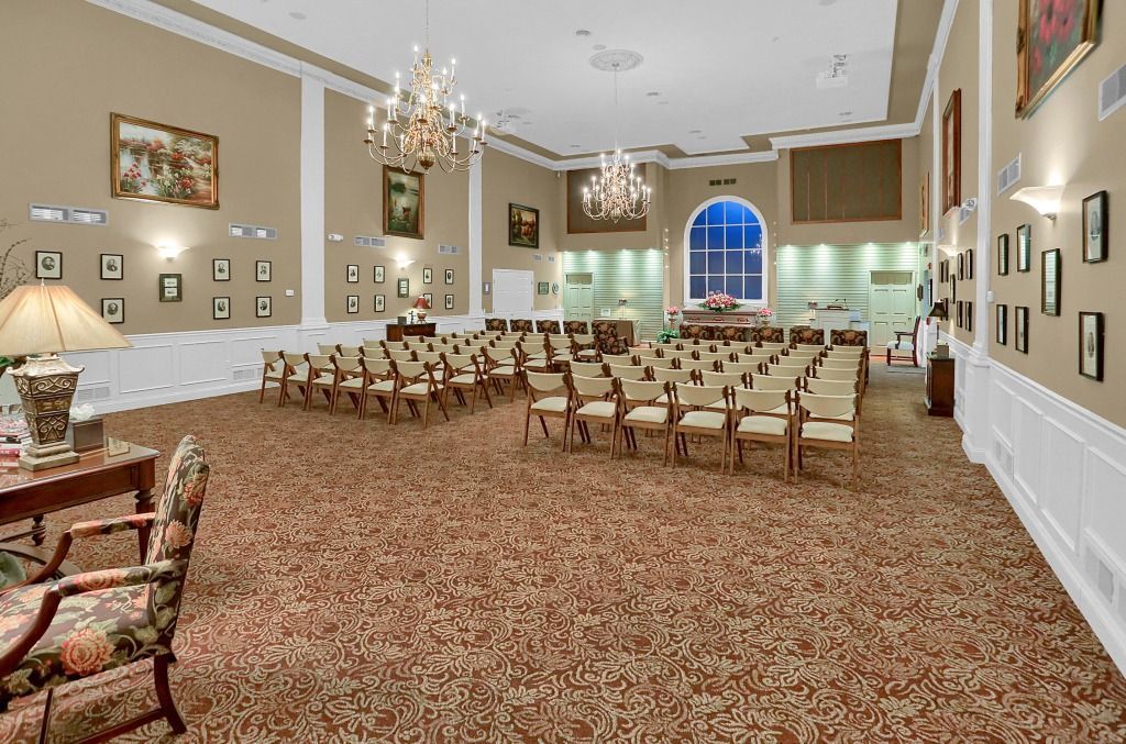 Large hall with rows of chairs, patterned carpet, chandeliers, and framed wall art.