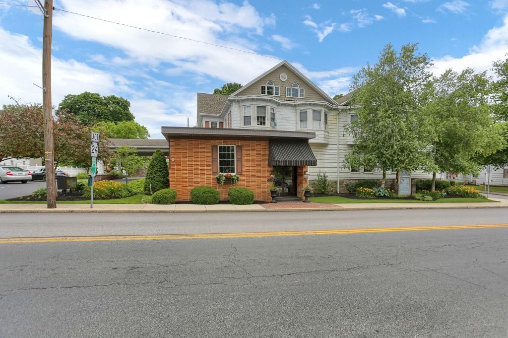 Street view of a small brick storefront with a gray house behind it and trees along the roadside