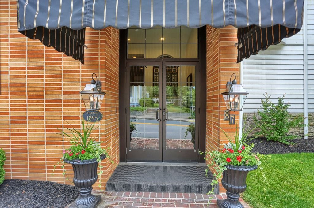 Front entrance with striped awning, brick walls, glass door, and potted plants beside the walkway.