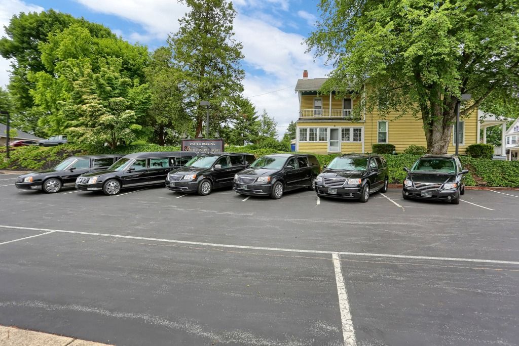 Black cars parked in a lot beside a yellow building and trees under a cloudy sky