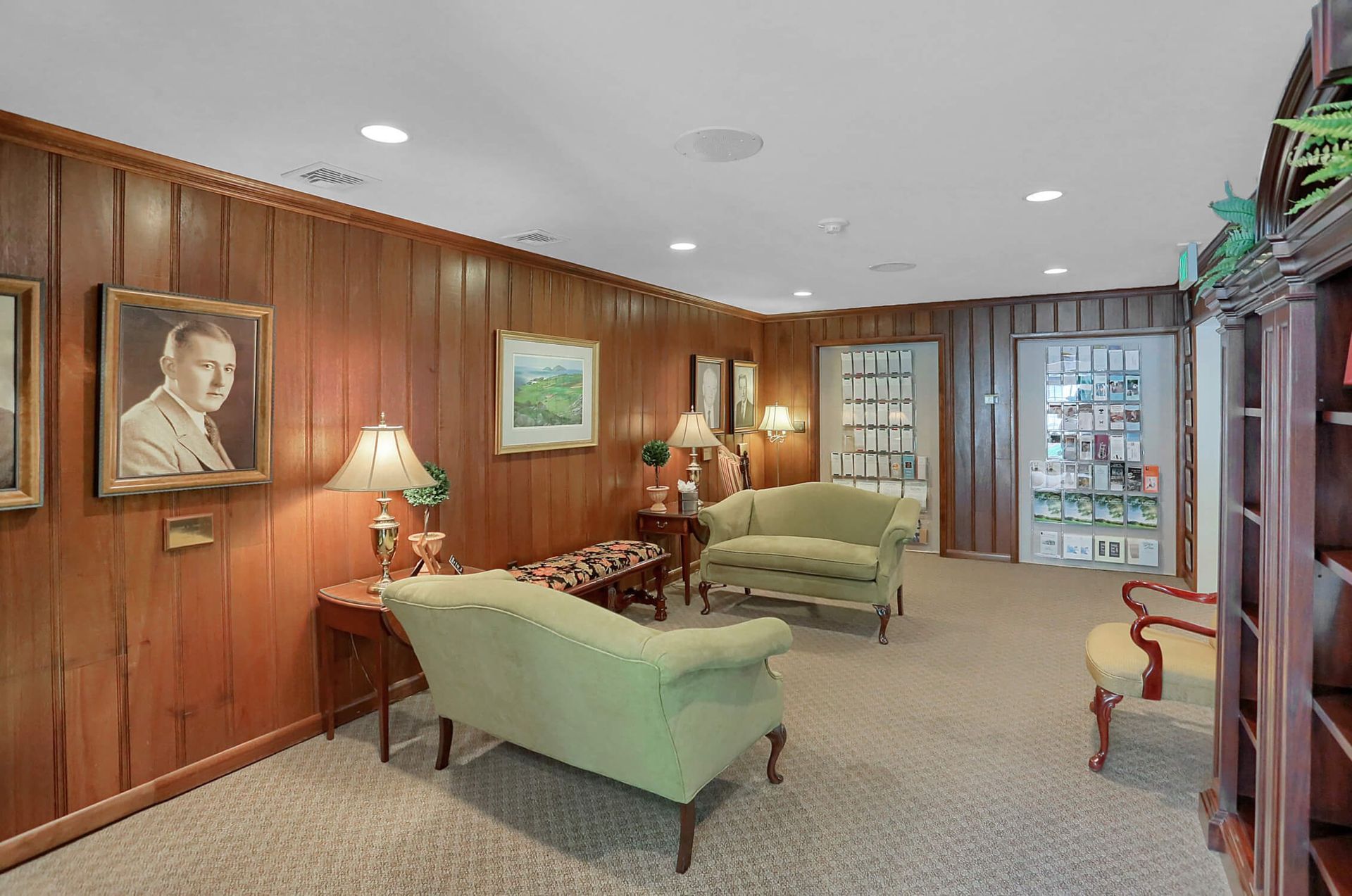 Wood-paneled lounge with green chairs, lamps, framed art, and glass doors in the back.