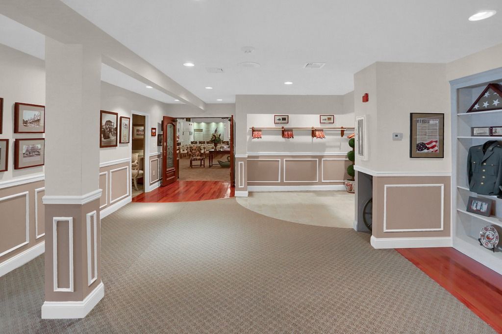 Bright lobby with carpeted hall, white walls, framed art, and a red-tiled area in the background