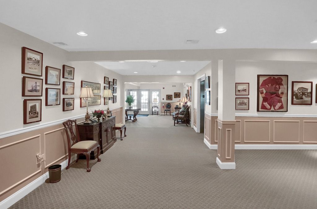 Bright hallway with framed art, white walls, beige wainscoting, and chairs along the left side.