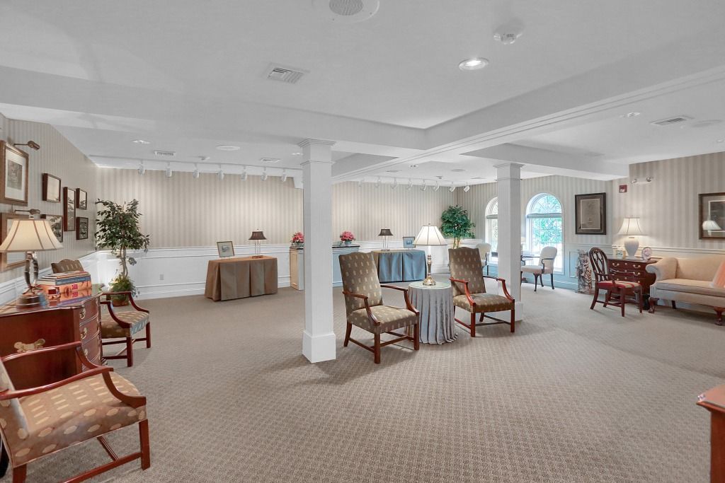 Bright, carpeted lounge with wicker chairs, small tables, plants, and a large arched window.