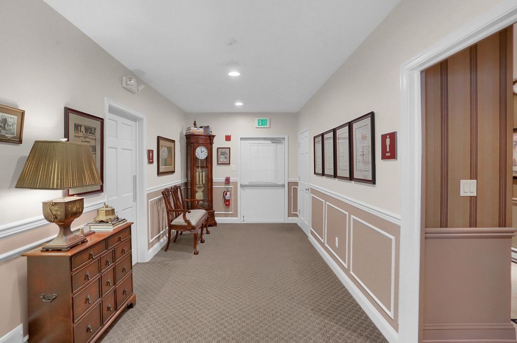 Bright hallway with carpet, white walls, framed art, wooden furniture, and a door at the end