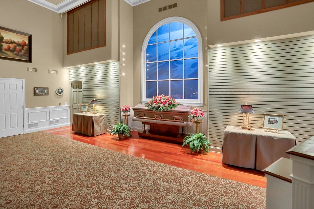 Decorated memorial room with flowers, a casket, and a large arched window at sunset