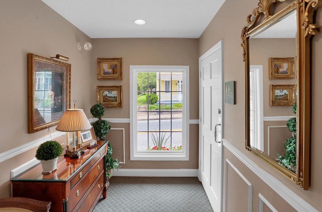 Bright hallway with beige walls, framed mirrors, a wooden console table, and a window looking onto a front yard.