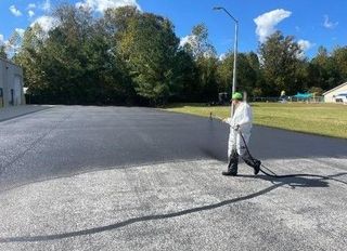 Worker spraying water on a paved road beside a grassy field and trees on a sunny day