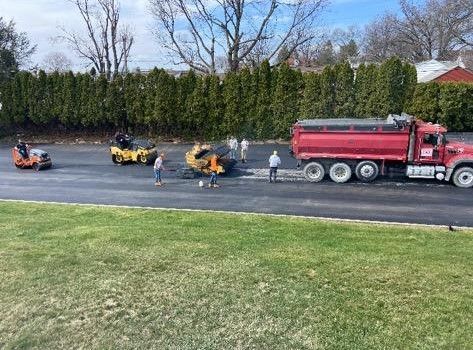 Workers spray pavement behind a red tanker truck and yellow equipment on a roadside.