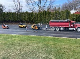 Workers spray pavement behind a red tanker truck and yellow equipment on a roadside.