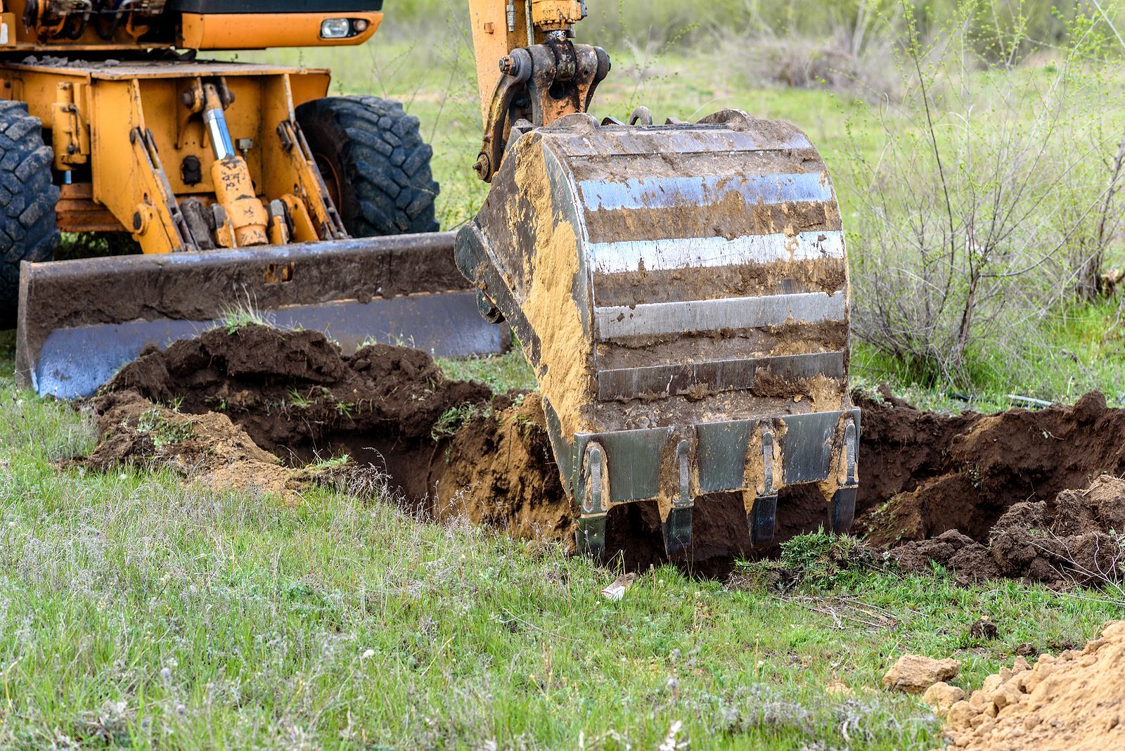 Yellow excavator digging in a grassy field, with soil piled beside the bucket