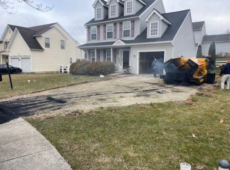 A driveway in front of a gray house is scorched, with a burned vehicle and firefighters at work.