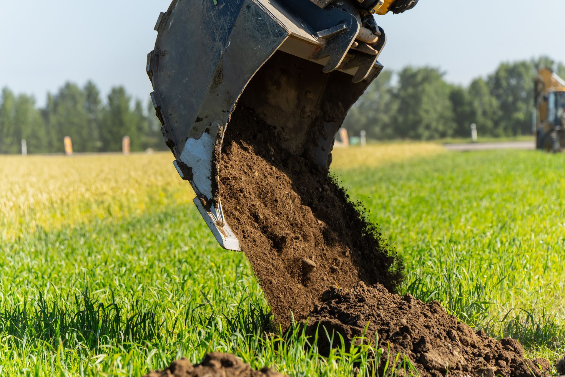 Excavator bucket dumping soil onto green grass in a field under a clear sky