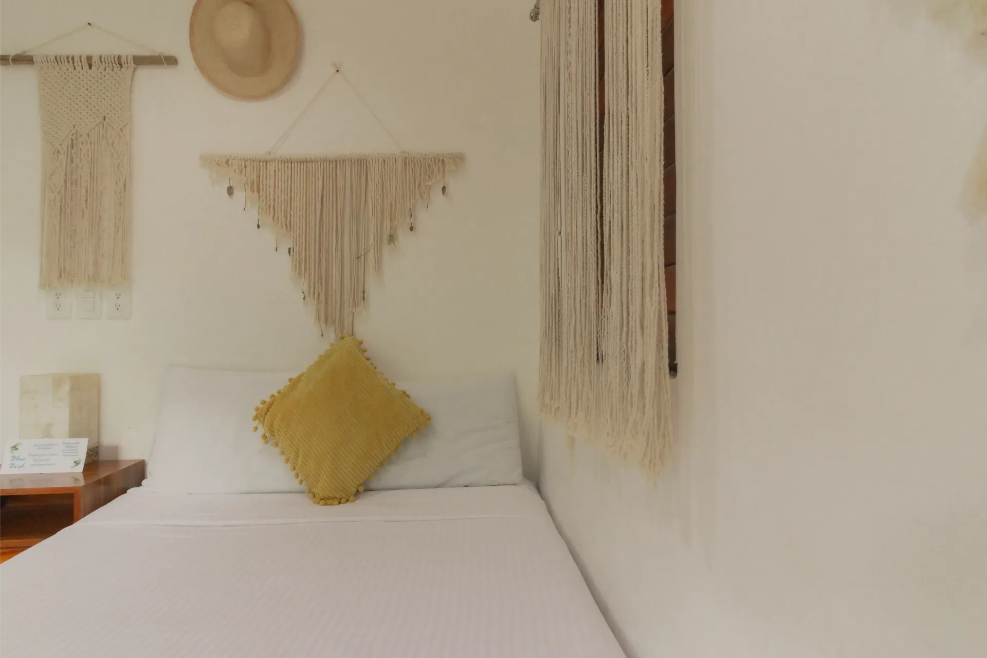 Dormitorio minimalista con cama blanca, tapices tejidos en las paredes y almohada amarilla con forma de diamante.