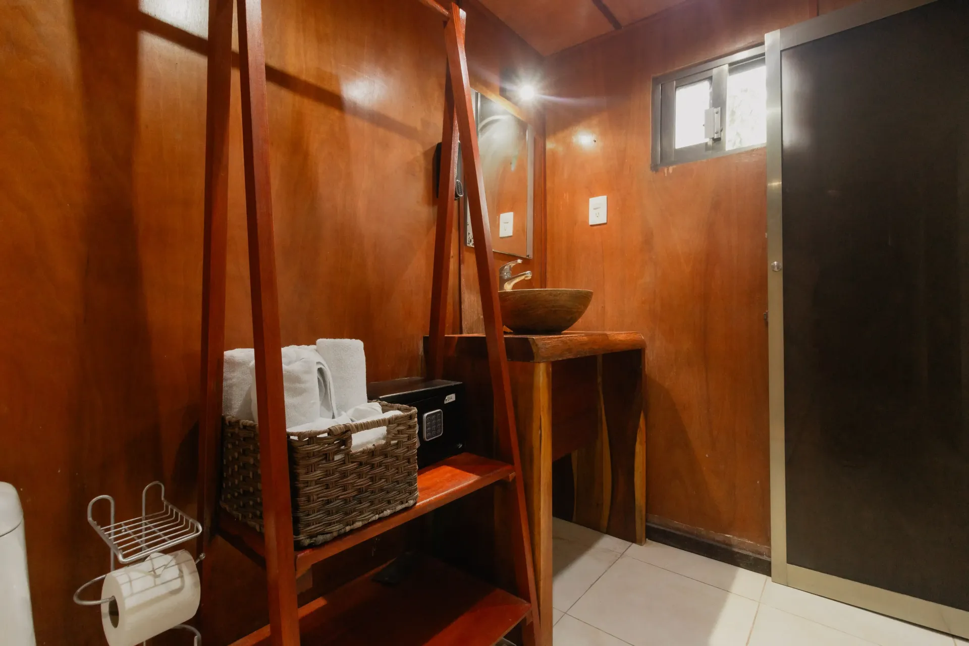 Baño revestido de madera con lavabo, espejo, inodoro, estanterías y suelo de baldosas.